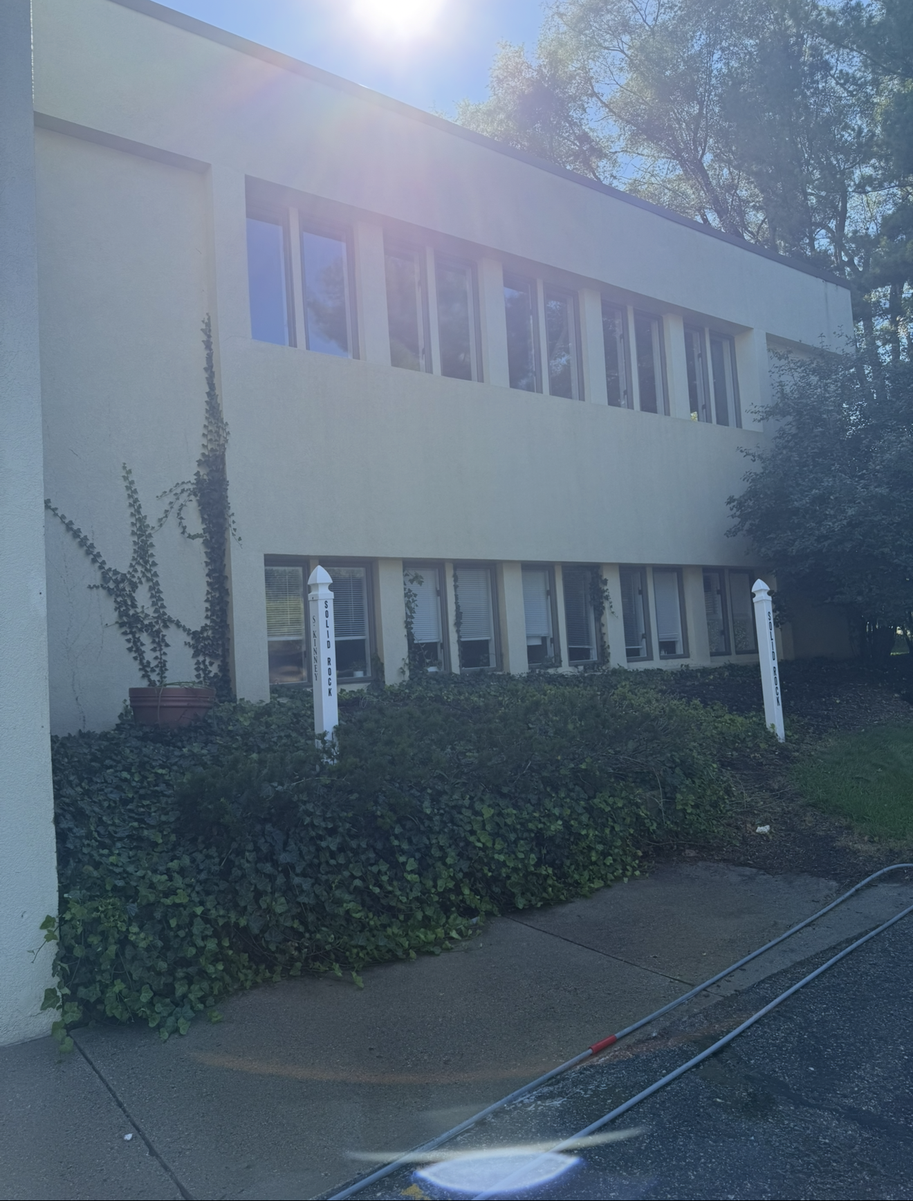 Sunlit two-story white building with ivy and hedges along a paved driveway