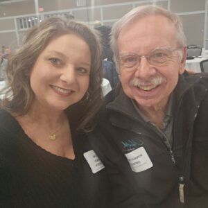 Two smiling people posing indoors at an event, wearing name tags