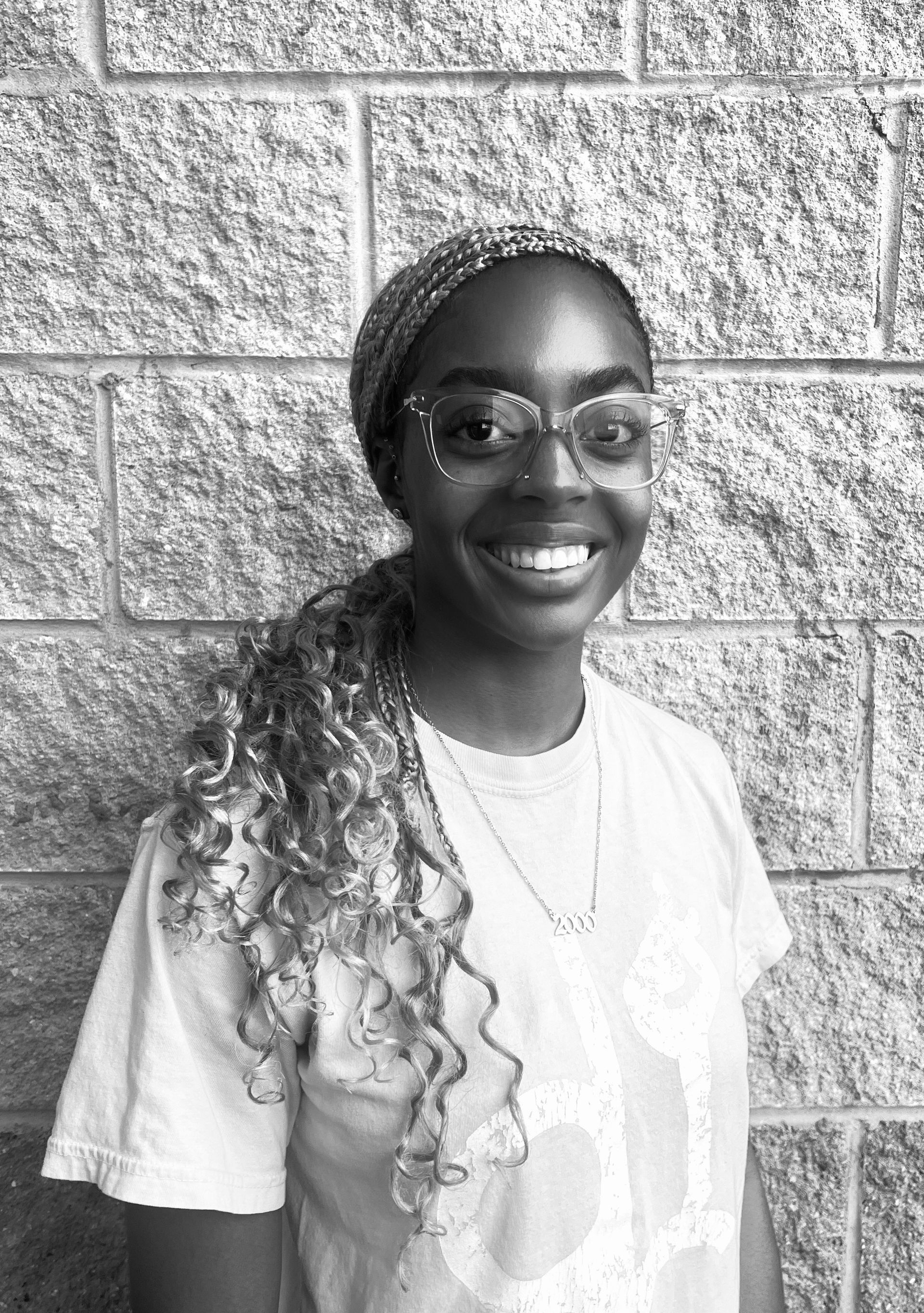 A woman wearing glasses and a headband is smiling in front of a brick wall.