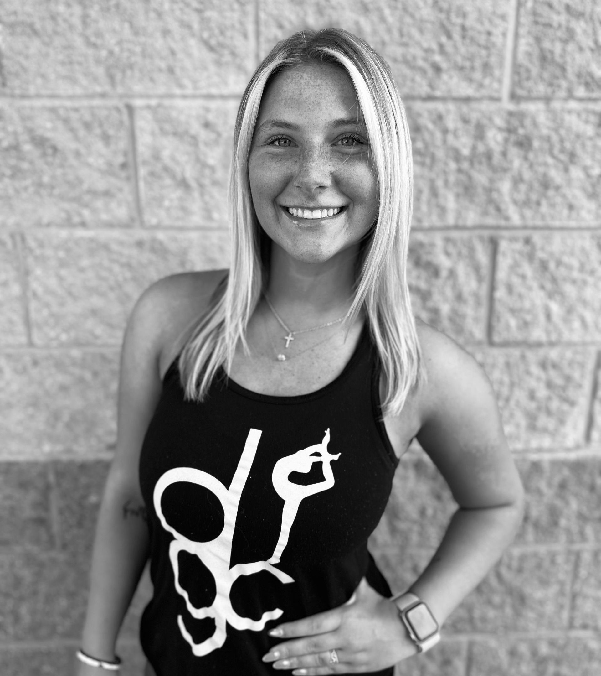 A woman in a black tank top is smiling in front of a brick wall.