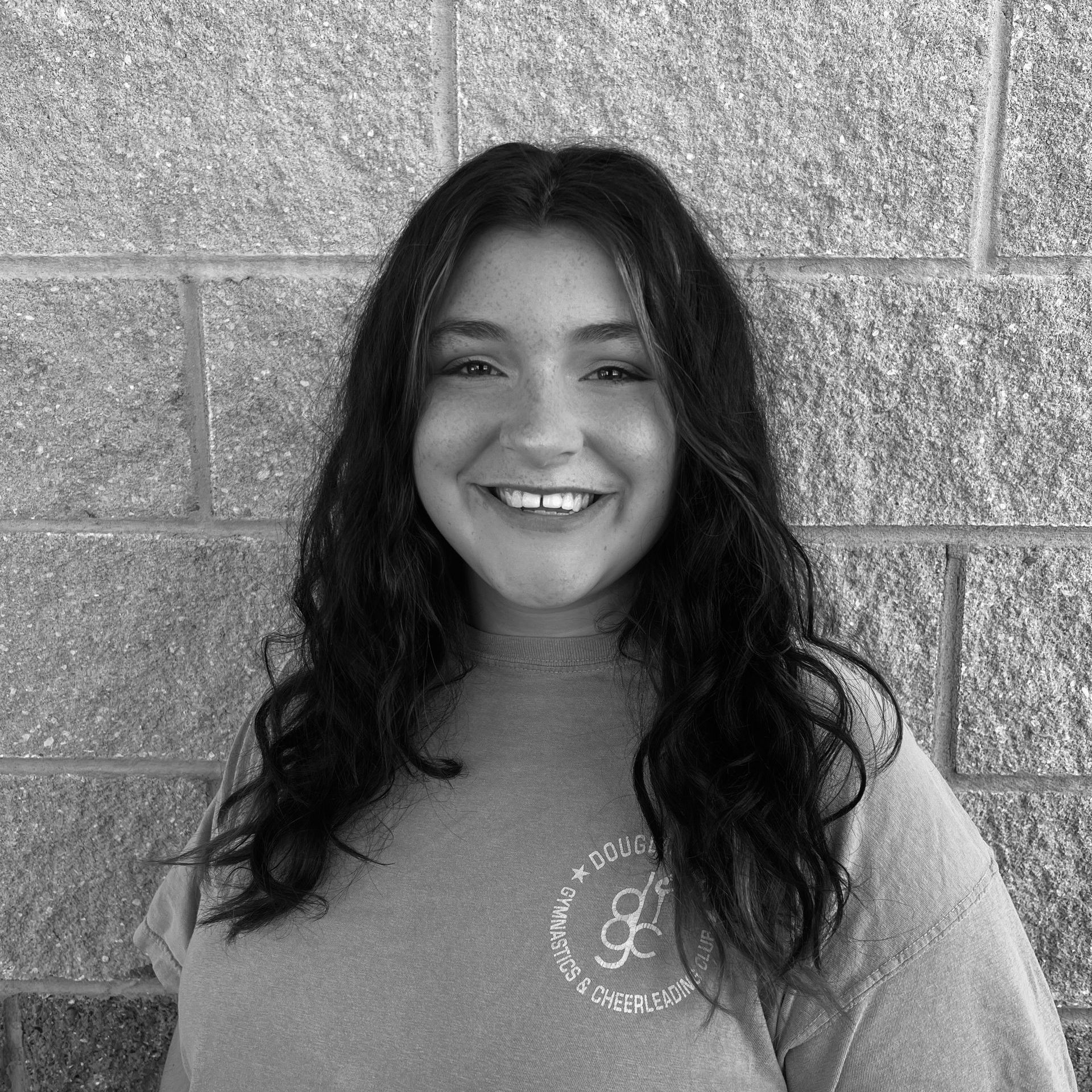 A black and white photo of a woman smiling in front of a brick wall.