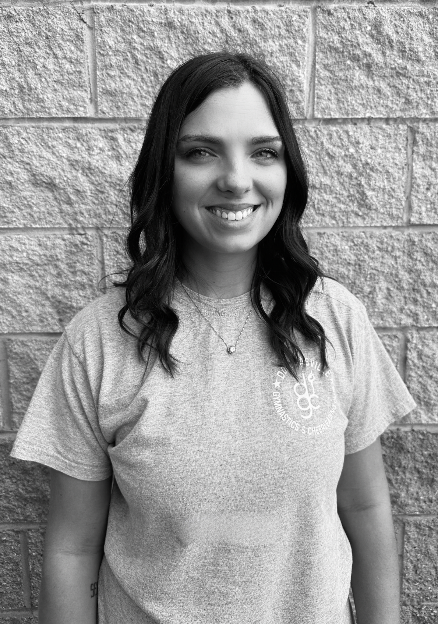 A woman in a grey t-shirt is smiling in front of a brick wall.