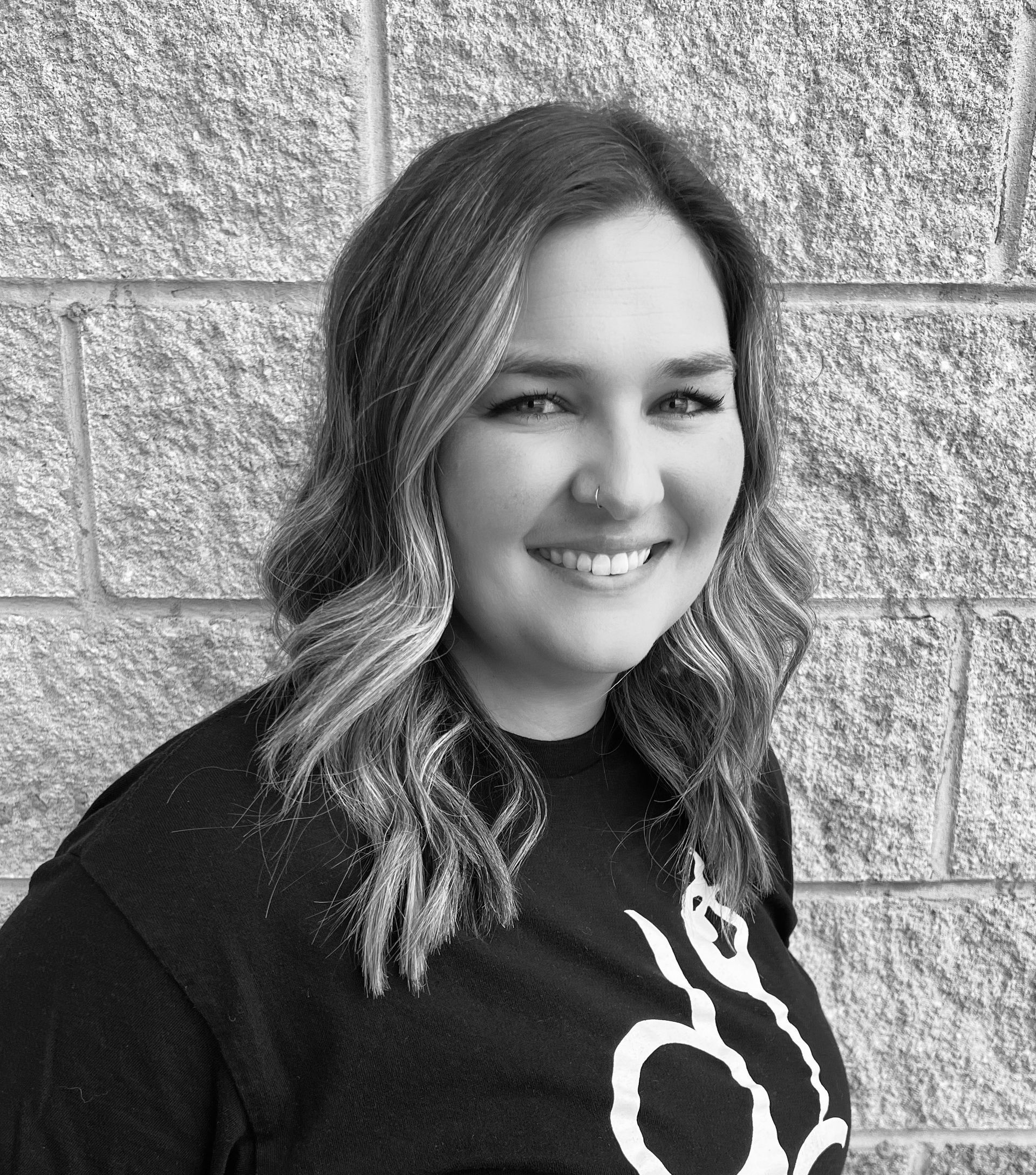 A black and white photo of a woman smiling in front of a brick wall.