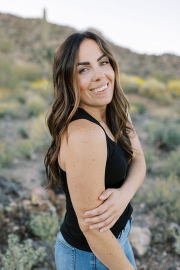 Smiling person with long, dark wavy hair wearing a black tank top, posed outdoors in a desert landscape at sunset.
