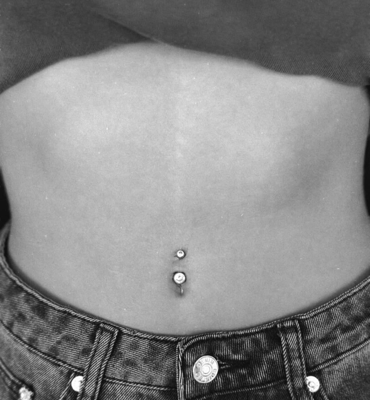 A black and white close-up of a navel piercing with two metal beads centered on the abdomen above denim jeans.