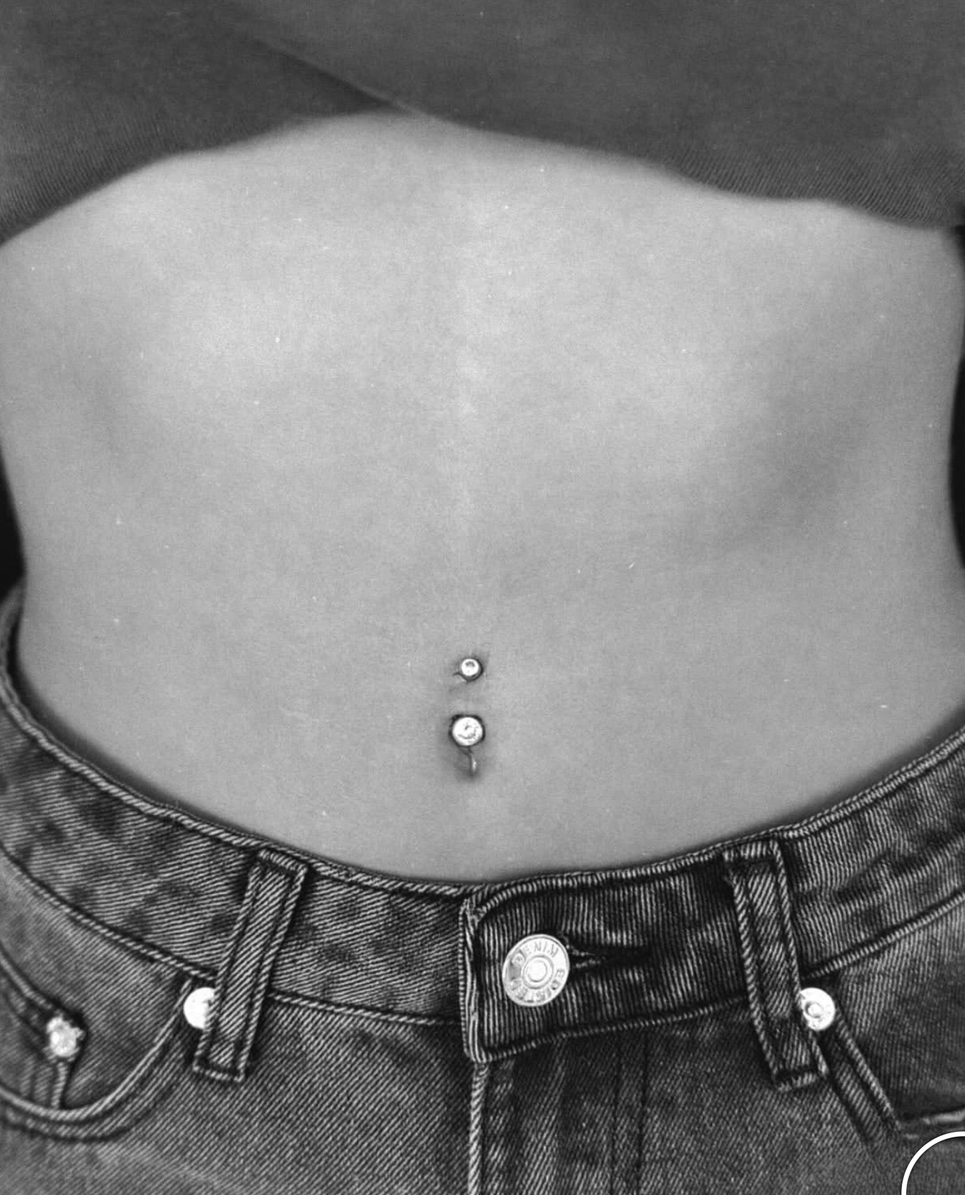 A close-up of a person's midriff wearing denim jeans, featuring a navel piercing with two small, clear gems.