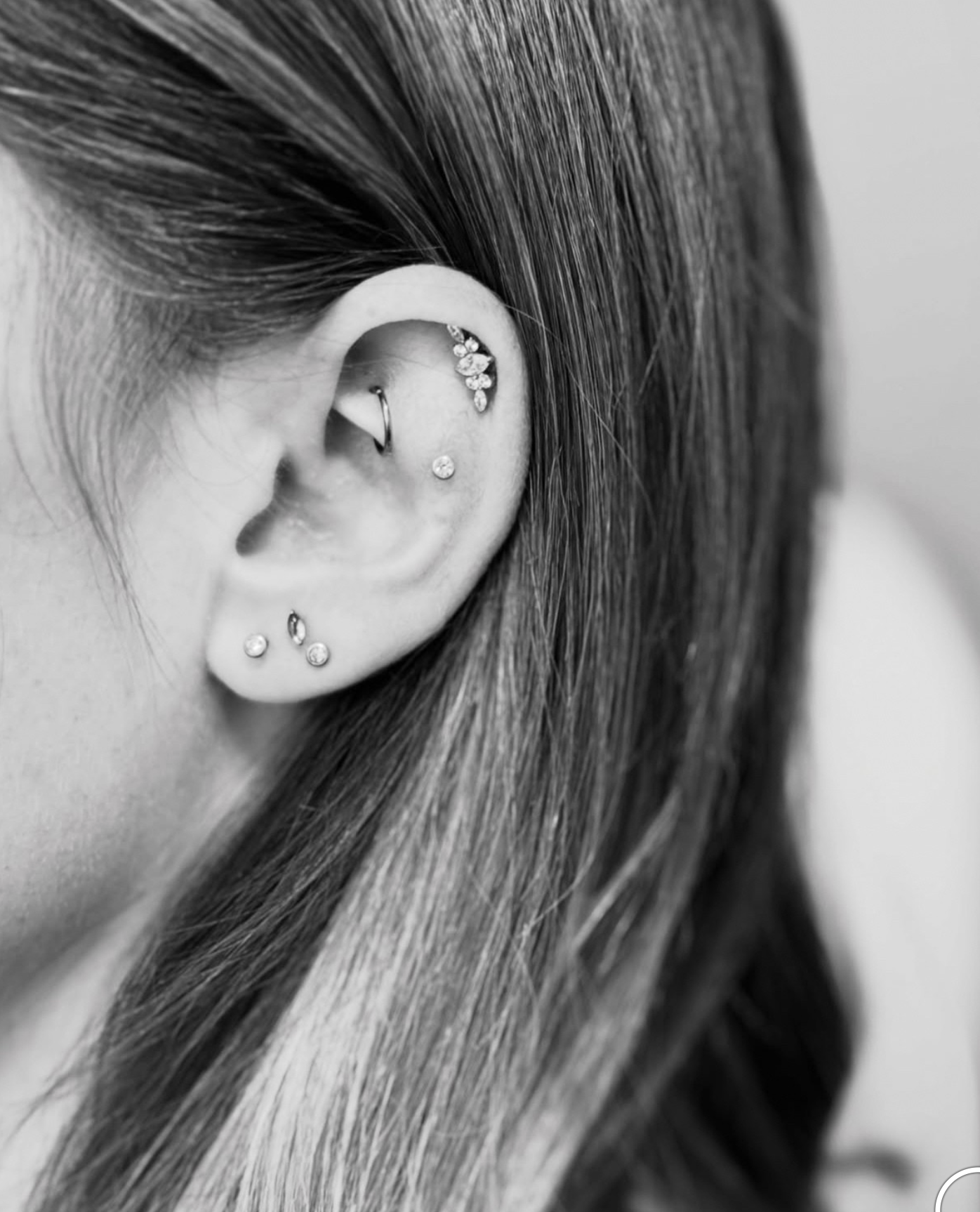Close-up, black-and-white photo of an ear with multiple piercings, including a rook, flat, and lobe studs.