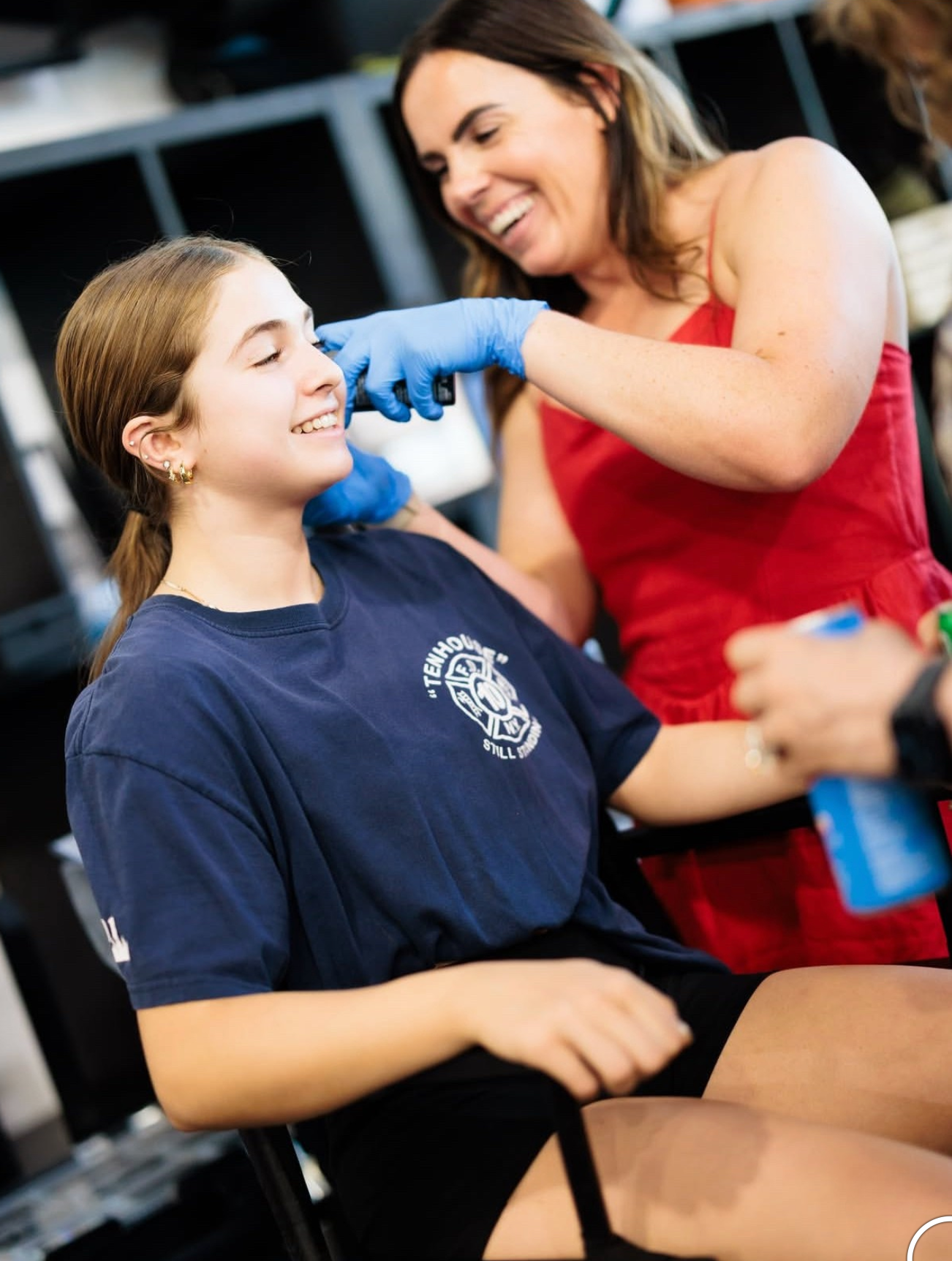 A person in a red dress wearing blue gloves performs an ear piercing on a person in a navy t-shirt.