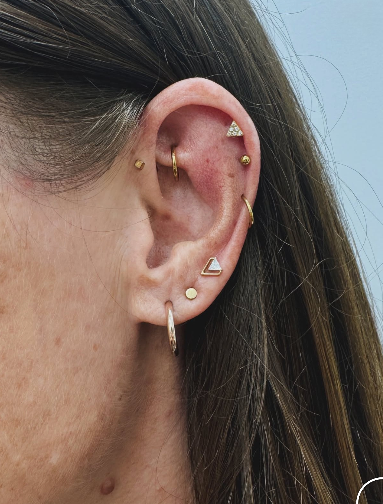 A close-up view of an ear featuring multiple gold piercings, including hoops, triangle studs, and small spherical beads.