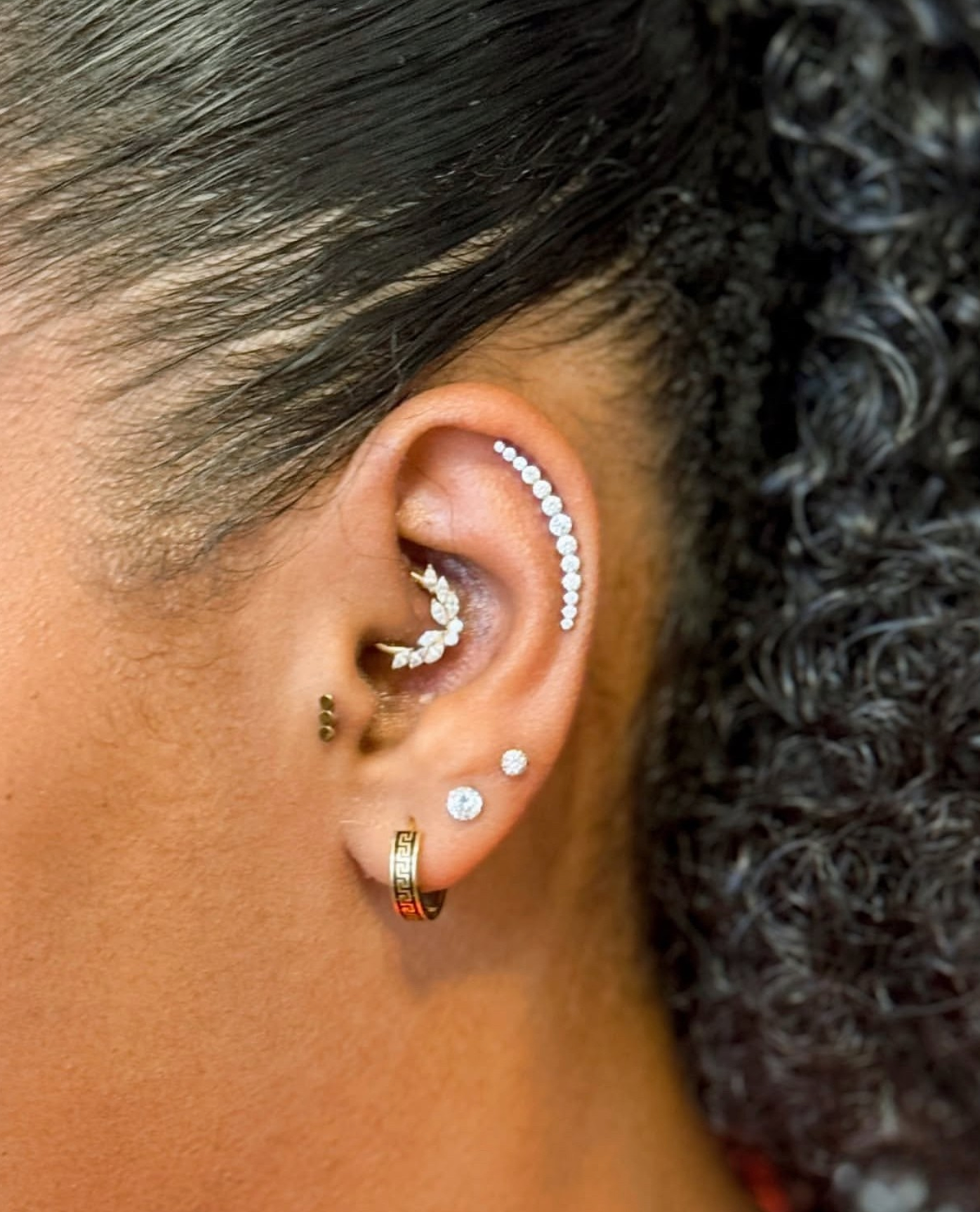 A person's ear featuring a curved, jeweled helix piercing, a leafy daith ring, and three lobe earrings.