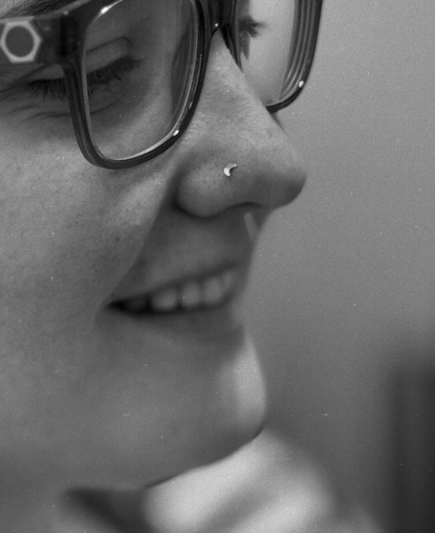 A close-up, black-and-white profile view of a smiling person wearing glasses and a small crescent moon nose stud.