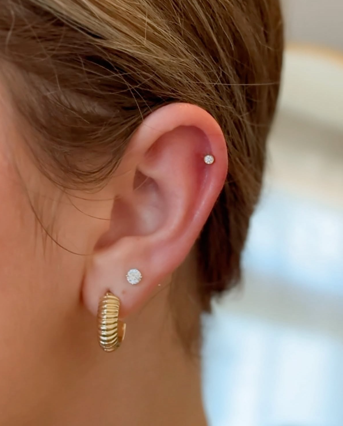 A close-up view of an ear with three piercings: a gold textured hoop, a small stud, and a cartilage stud.