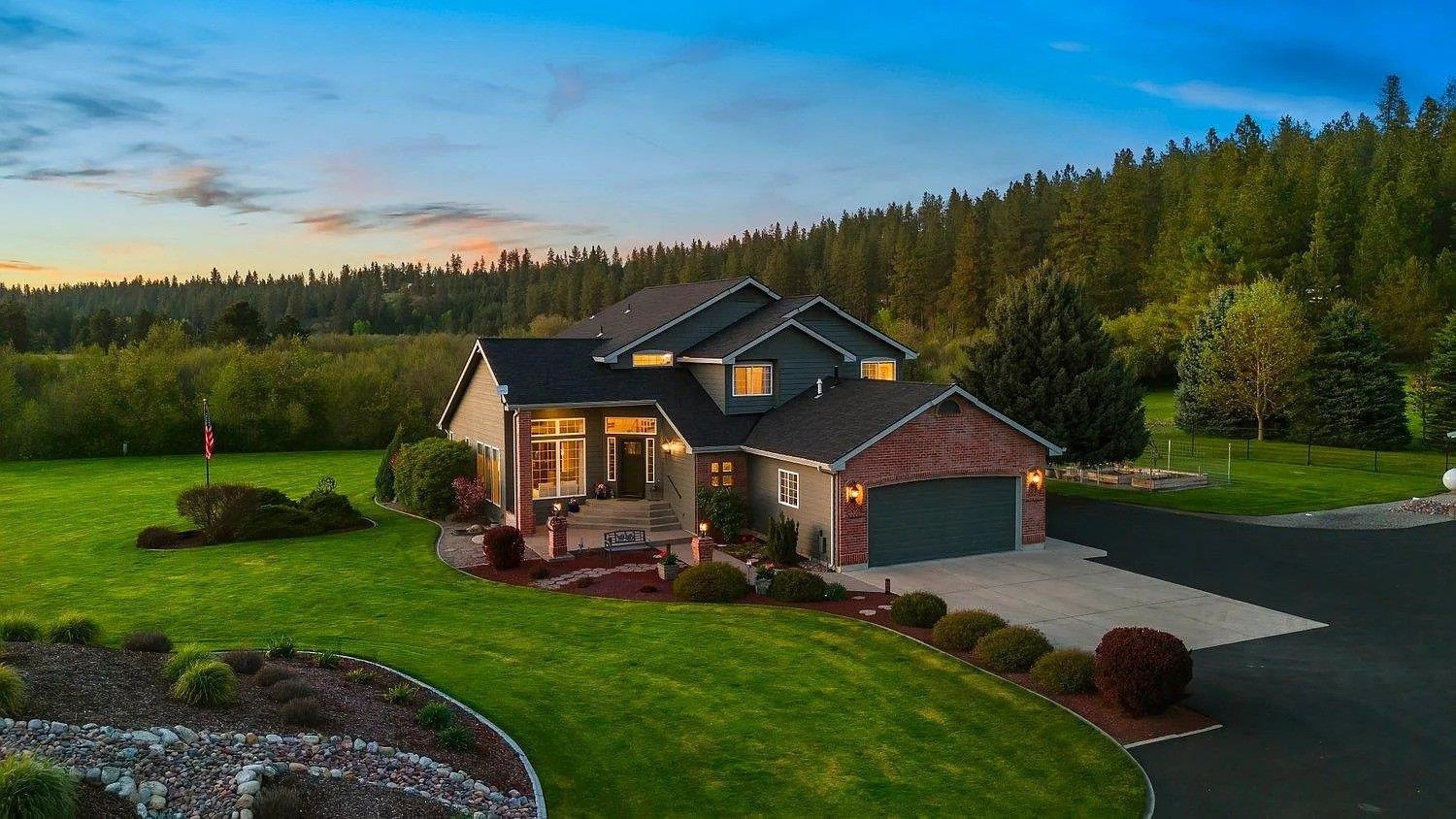 Modern house with lit porch at dusk, set on a green lawn beside a forested road.