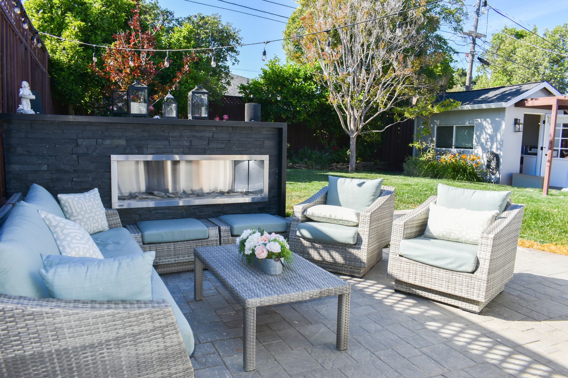 Outdoor patio with wicker seating, coffee table, and a built-in fireplace in a sunny backyard.