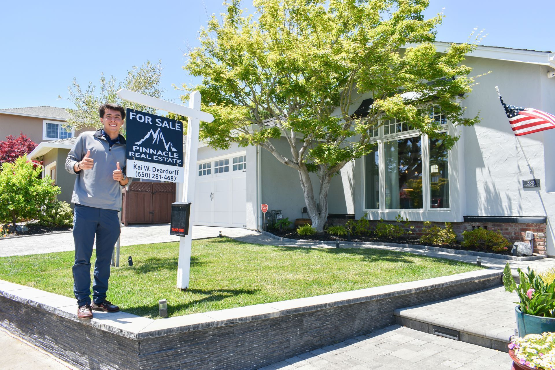 Man standing outside a white house beside a real estate sign, with an American flag and tree in front
