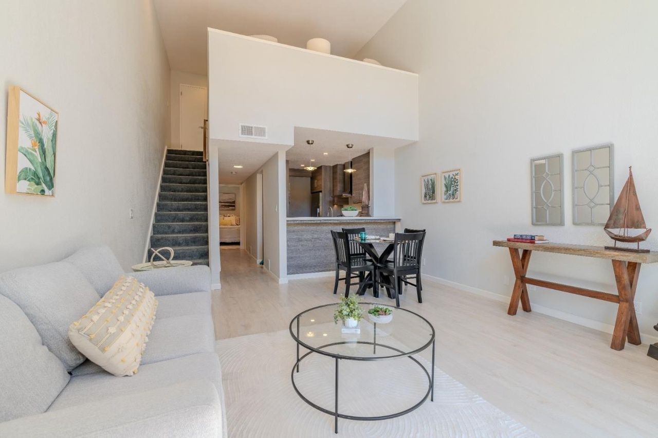 Bright, open living room with white walls, gray sofa, glass coffee table, stairs, and small dining area