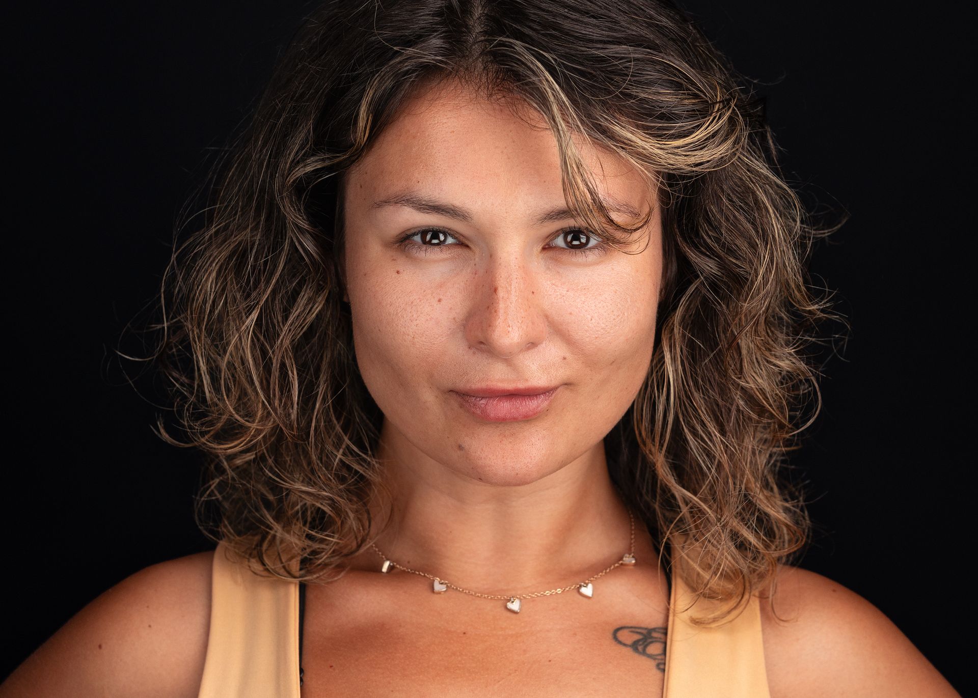 A person with wavy brown hair looks directly at the camera against a plain black background, wearing a simple necklace.