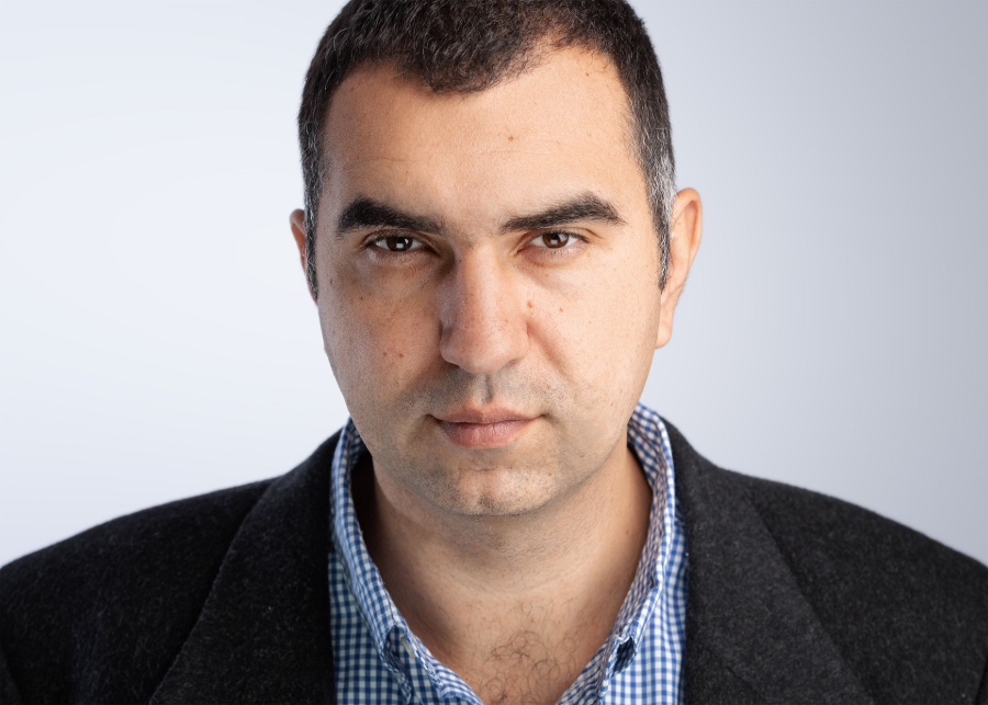 Man in dark blazer and blue checkered shirt, facing forward, serious expression.