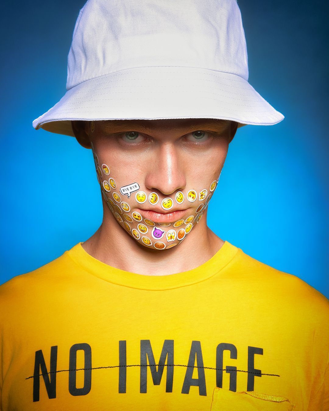 Man wearing a bucket hat and yellow shirt with emojis on his face against a blue backdrop.