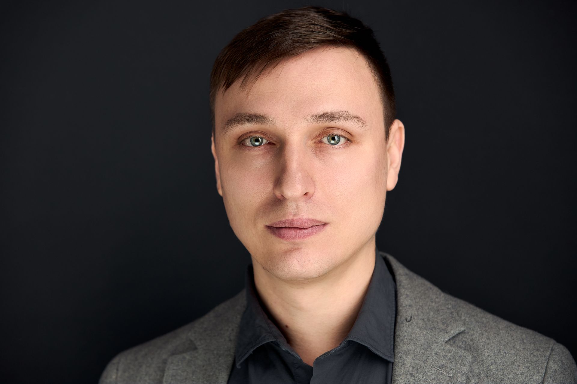 Man in gray blazer, dark shirt, looks directly at the camera, dark background.