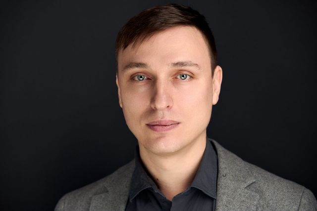 Man with light skin wearing a grey blazer and dark shirt, looking at the viewer.