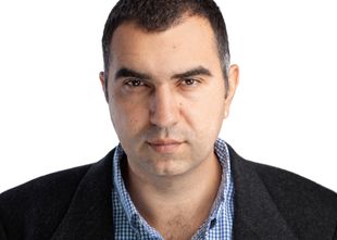 Man with dark hair and a serious expression, wearing a blue plaid shirt and a dark blazer.