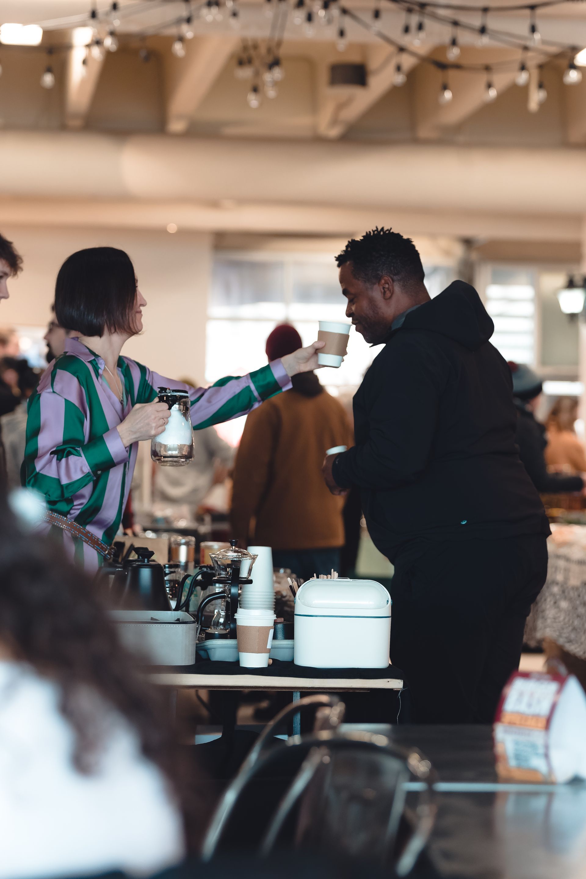 A person in a green and purple striped shirt hands a coffee cup to a person in a black hoodie at an indoor market stall.