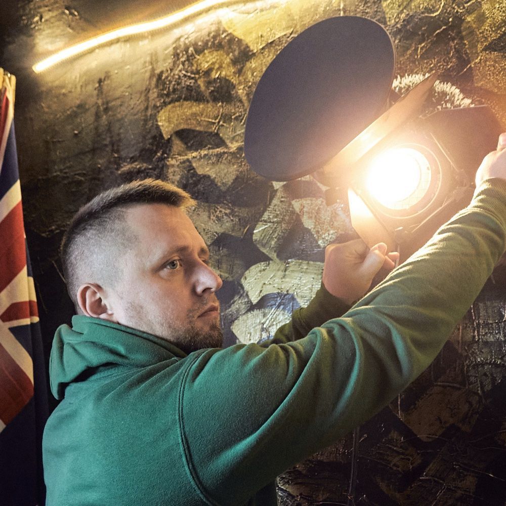 Man in green hoodie adjusts a spotlight on a textured wall; Union Jack flag visible.