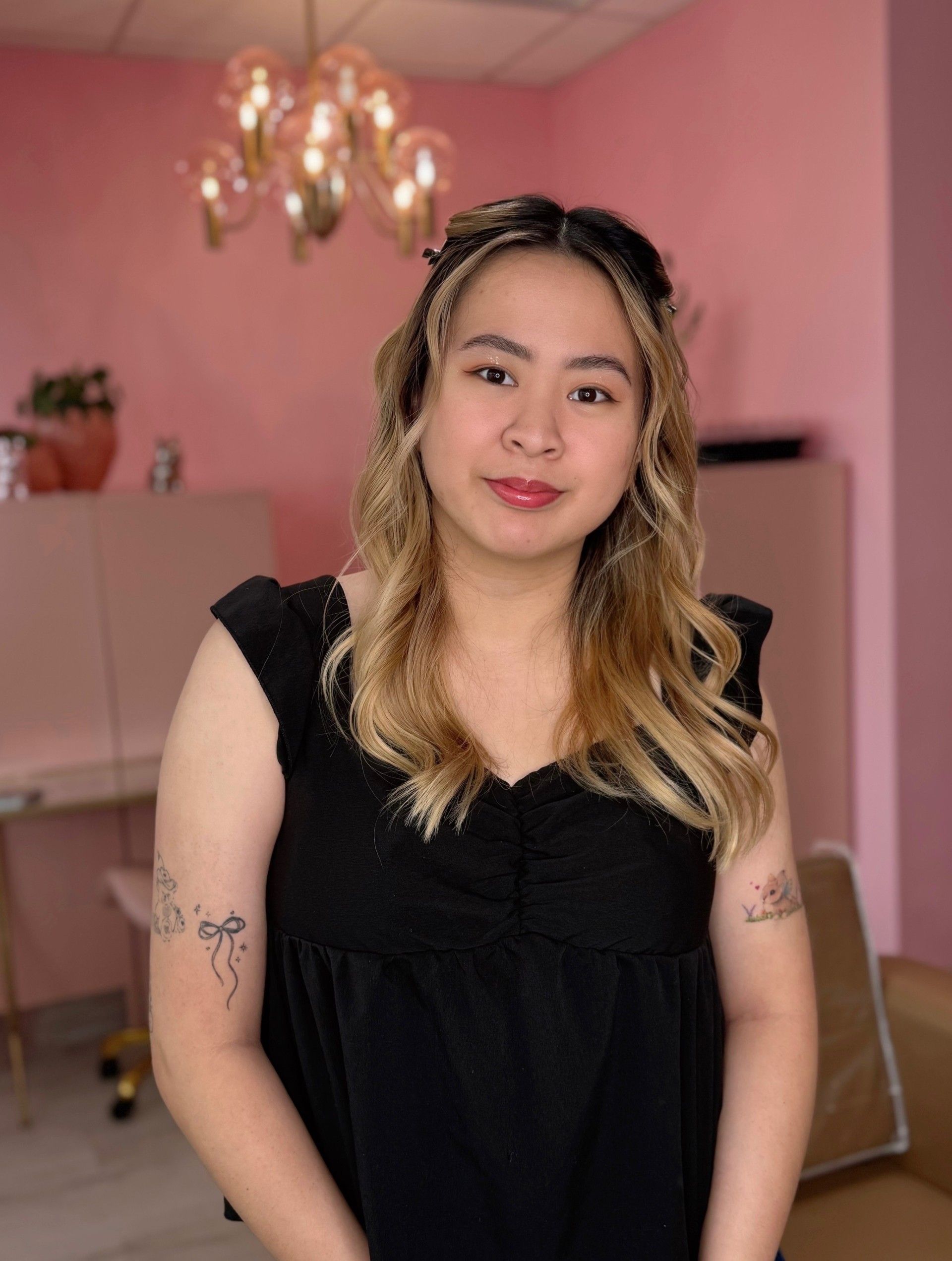 A person with wavy blonde-highlighted hair in a black top stands in a pink room under a glass chandelier.
