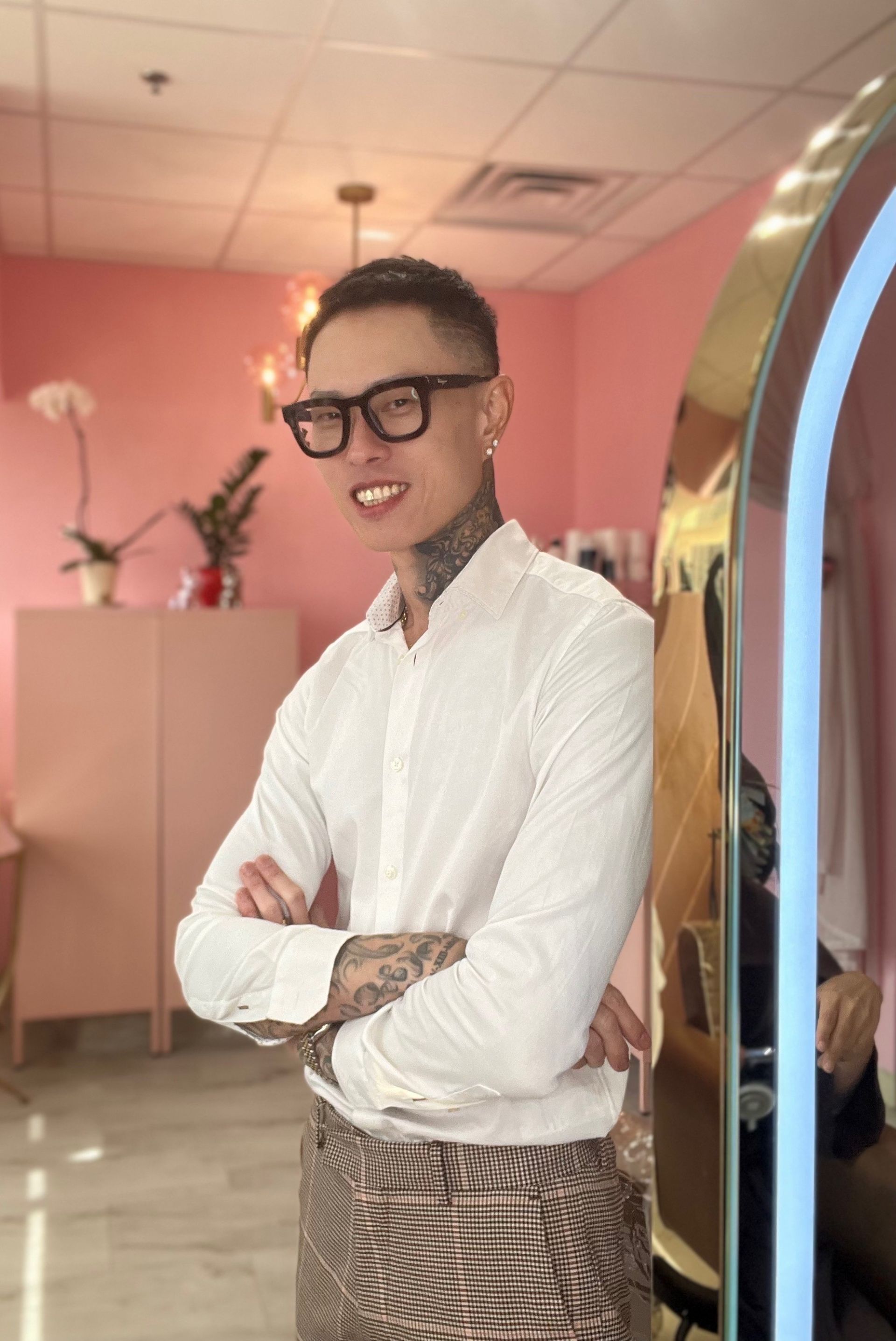 A person wearing glasses, a white collared shirt, and patterned pants standing with arms crossed in a pink room.