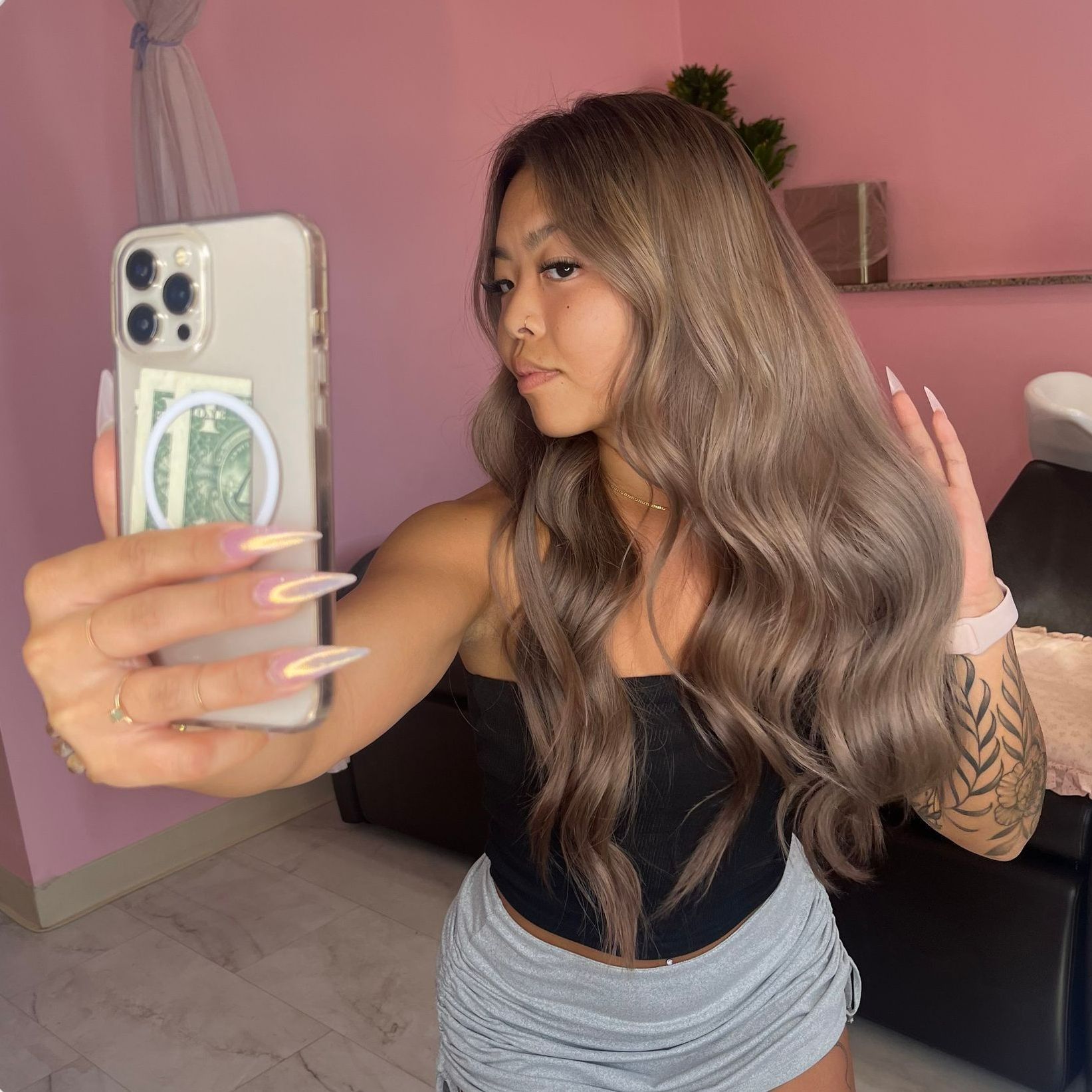 A person with long, wavy brown hair takes a mirror selfie, wearing a black top, gray skirt, and rings, in a pink room.