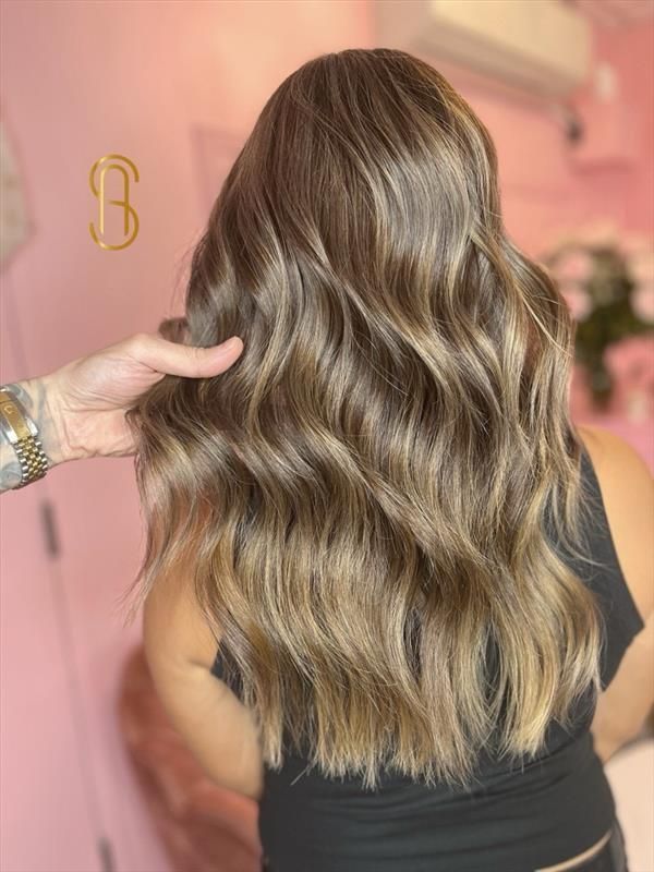 Long, wavy brown hair with blonde highlights styled in a salon with a pink interior background.