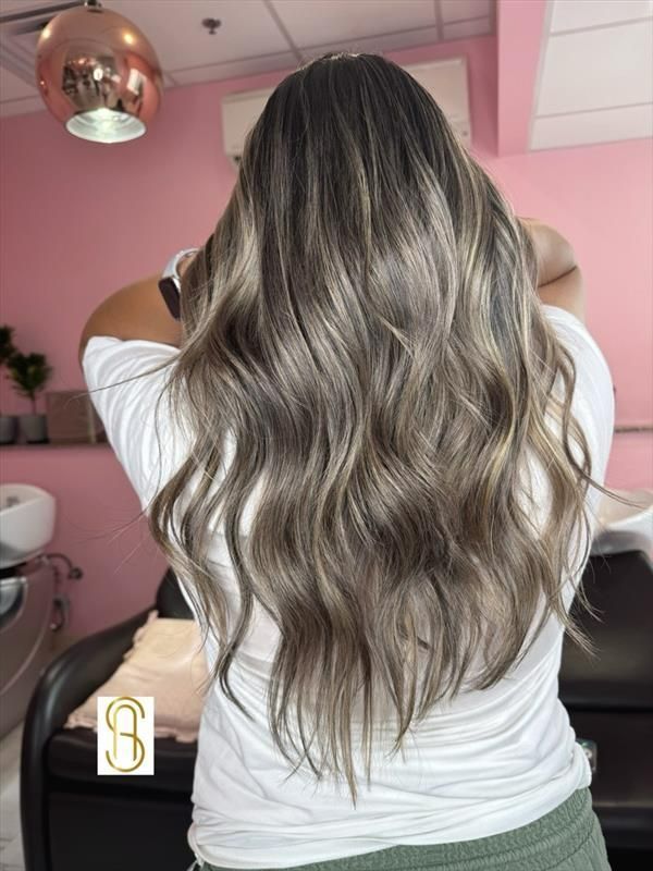 A person with long, wavy, ash-brown highlighted hair seen from the back in a hair salon.