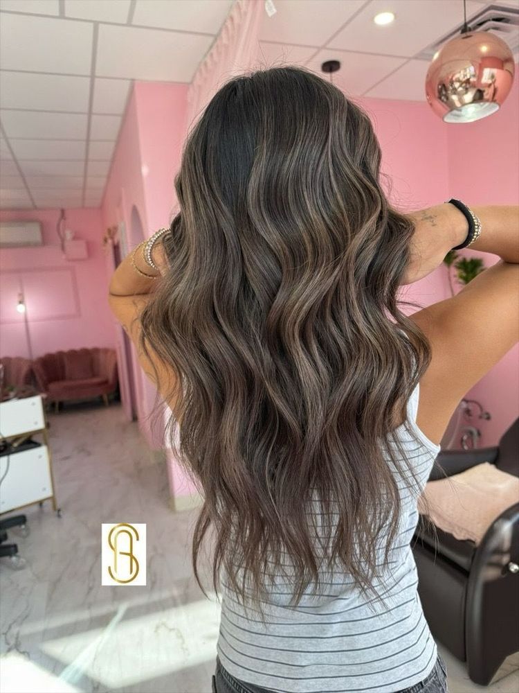 A person with long, wavy, brunette hair with blonde highlights stands in a pink-themed hair salon.