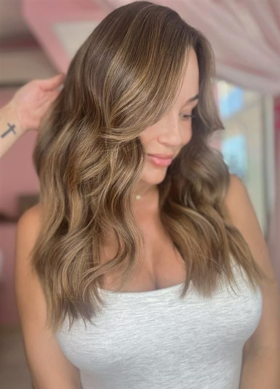 A person with wavy, highlighted light-brown hair, wearing a gray tank top, poses in front of a soft pink backdrop.