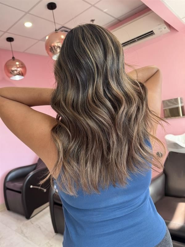 Back view of someone with wavy, highlighted light brown hair wearing a blue tank top inside a salon with pink walls.