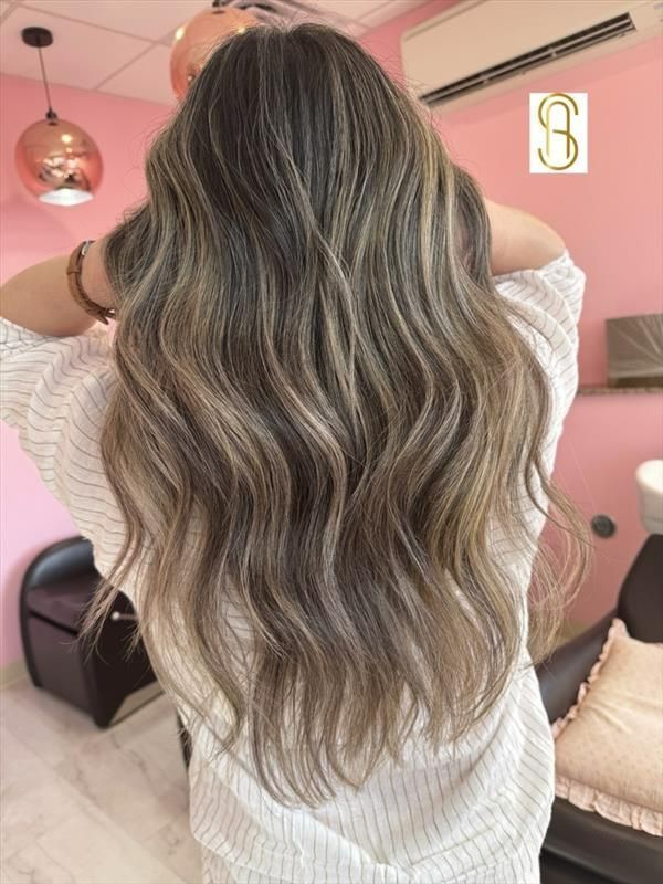 A person with long, wavy, ash-brown hair featuring blonde highlights, seen from behind in a salon with pink walls.