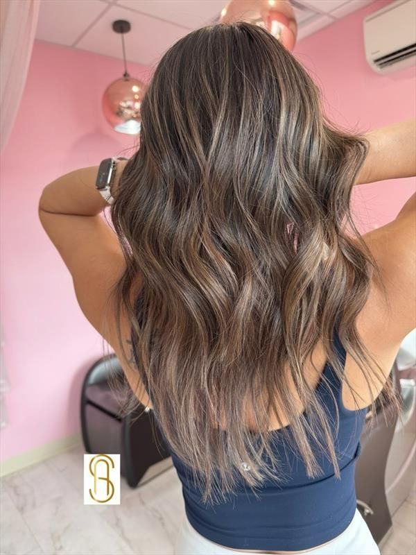 A person with long, wavy brown hair with blonde highlights, shown from the back in a hair salon.