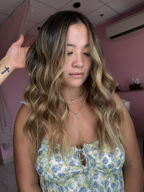 A person with long, wavy, highlighted brunette-to-blonde hair stands in a salon with pink walls.