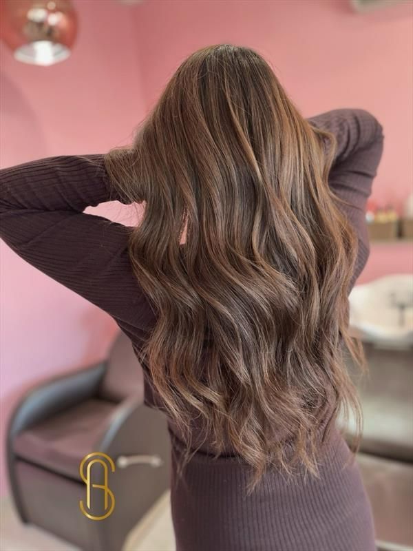 A person with long, wavy, light brown hair with highlights, seen from behind in a salon setting.