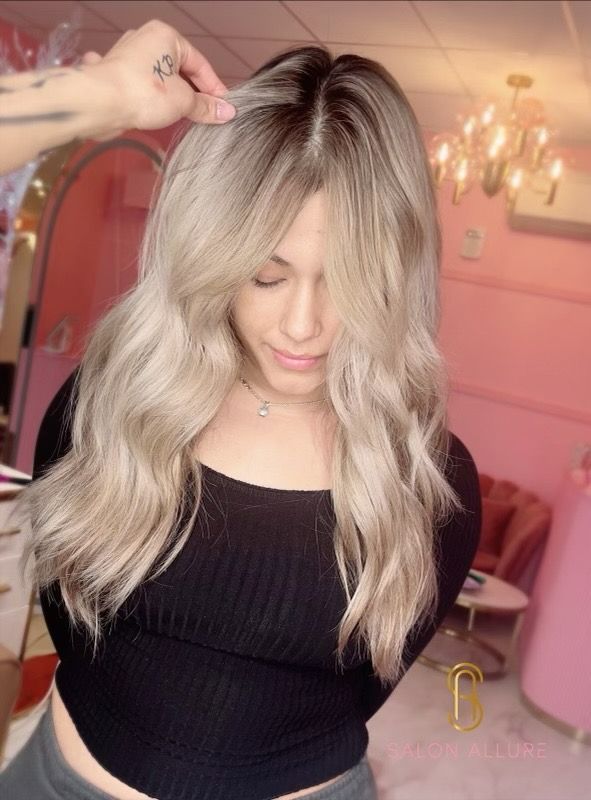 A hairstylist pulls back a section of a client's long, wavy, cool-toned blonde hair, showing a dark root smudge.