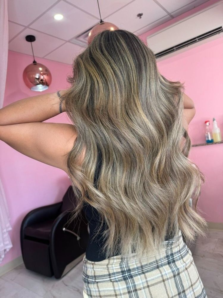 Long, wavy hair with blonde highlights styled in a salon with pink walls and pendant lights.
