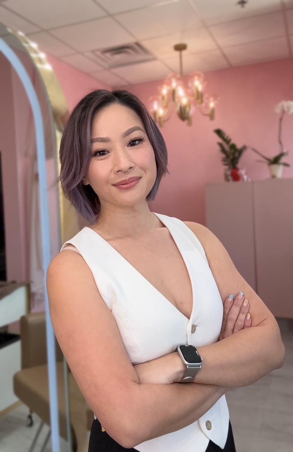 A person with short, lavender-tinted hair wearing a white vest and watch, posing with crossed arms in a pink room.
