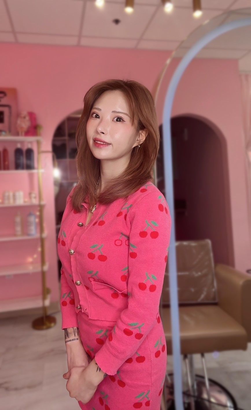 A person with shoulder-length brown hair wears a bright pink cherry-patterned cardigan in a pink-themed salon.