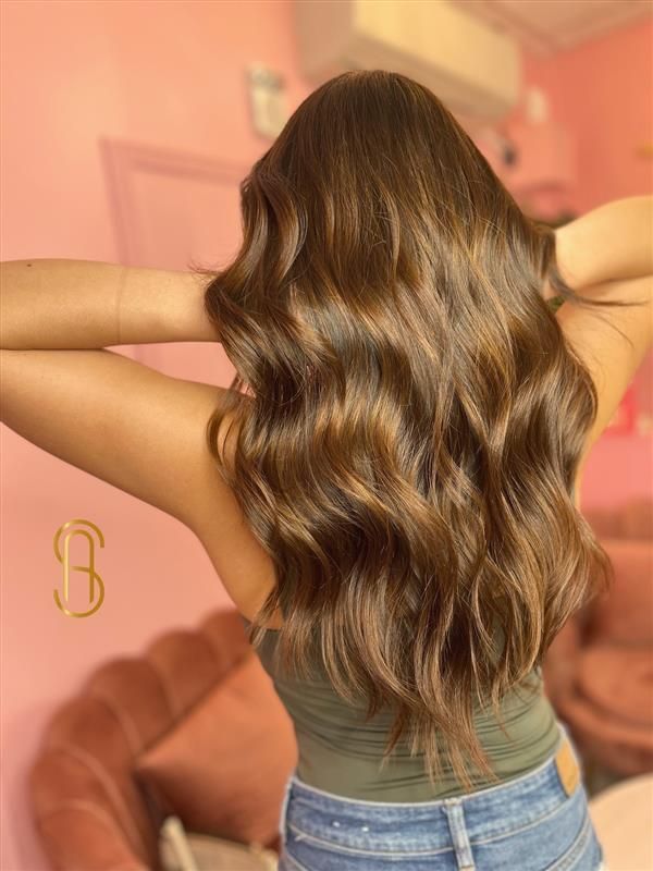 Long, wavy brown hair viewed from behind, worn by a person in a green tank top and jeans against a pink background.