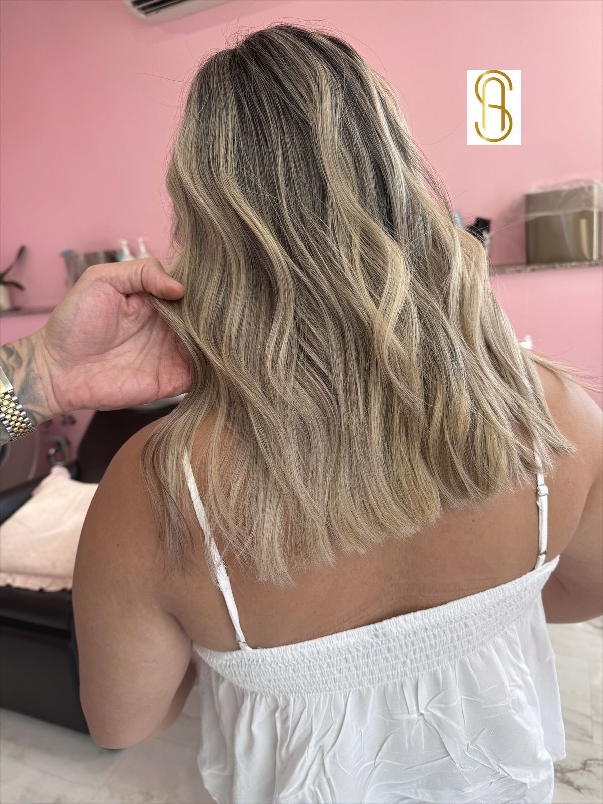 A stylist’s hand holds up a section of shoulder-length, highlighted blonde hair with soft waves in a salon setting.