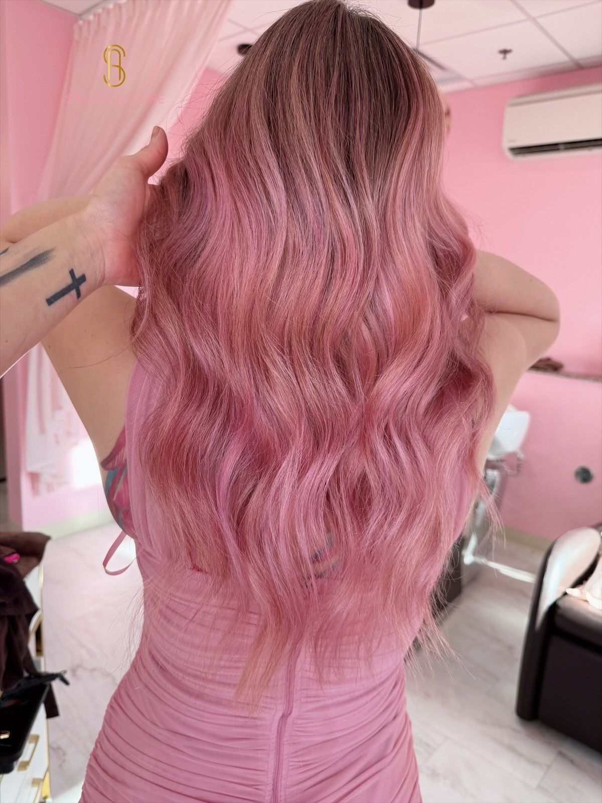 A person from behind displays long, wavy, rose-gold hair, wearing a matching pink dress in a salon setting.