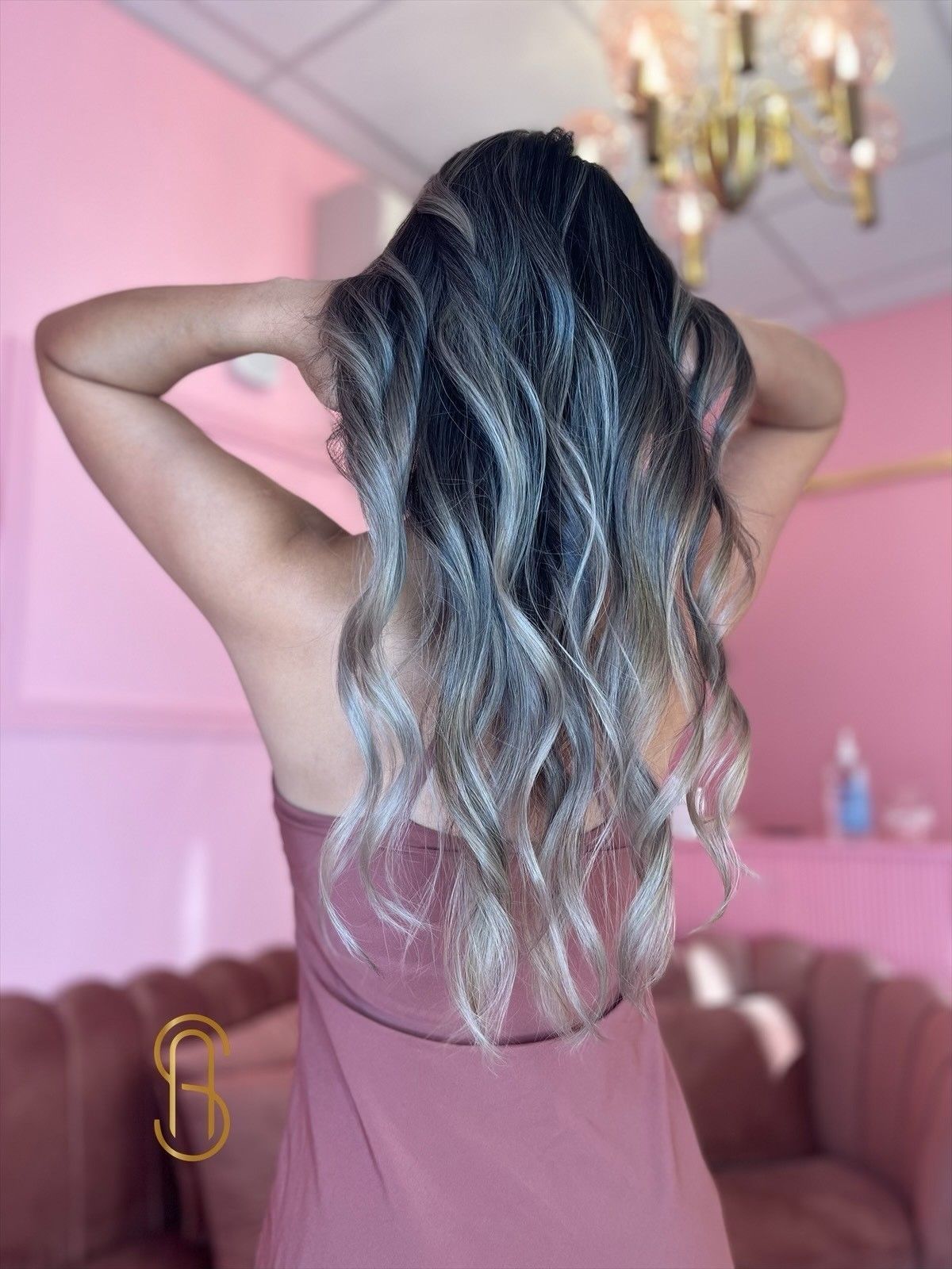 A person with long, wavy hair featuring dark roots and ash-blonde highlights, seen from behind in a pink-themed room.