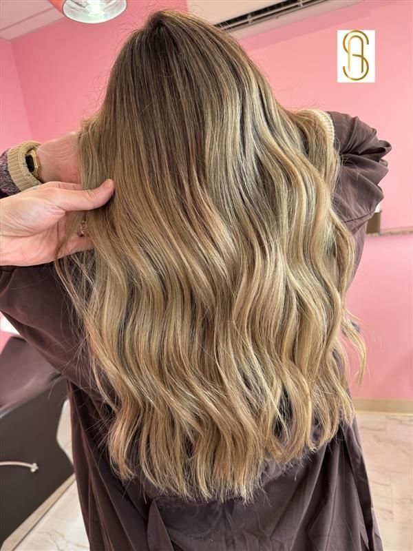 Long, wavy hair with multi-tonal blonde highlights and a darker root, shown from behind in a salon setting.