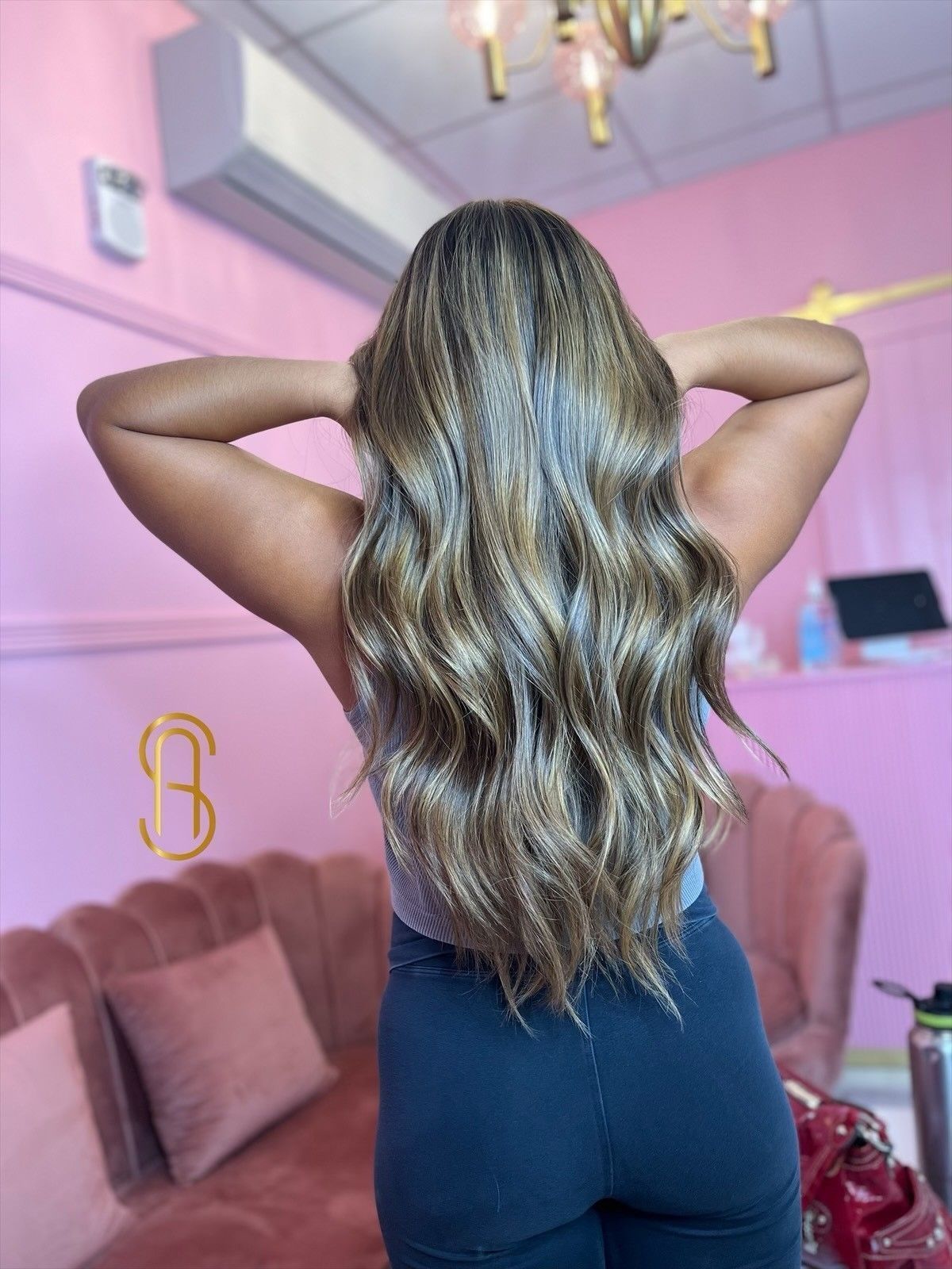 A person seen from behind in a pink salon, showcasing long, wavy, highlighted brown hair with their hands on their head.